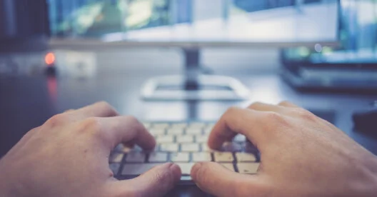 Hands are typing on a white keyboard, screen in the blurry background
